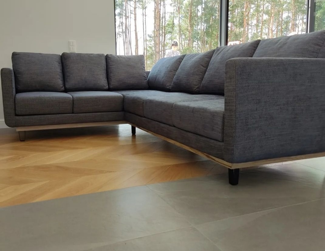 Szary, narożny, tapicerowany zestaw wypoczynkowy z poduszkami, na drewnianej ramie, z niskim oparciem, na tle okna z widokiem na las i parkiet w jodełkę.