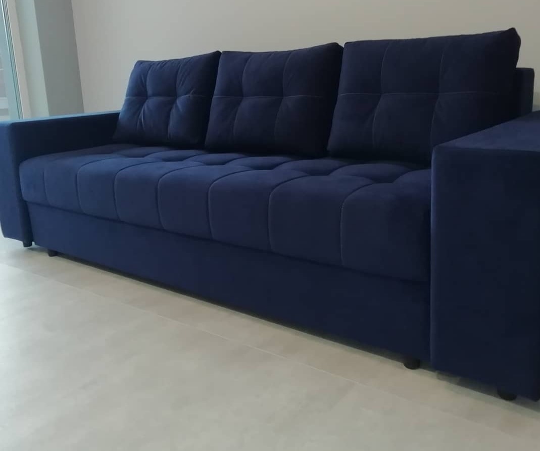 Granatowa sofa z tapicerowanymi kwadratami, ustawiona na jasnej podłodze. Minimalistyczny design, widoczne przeszycia i miękka faktura materiału.