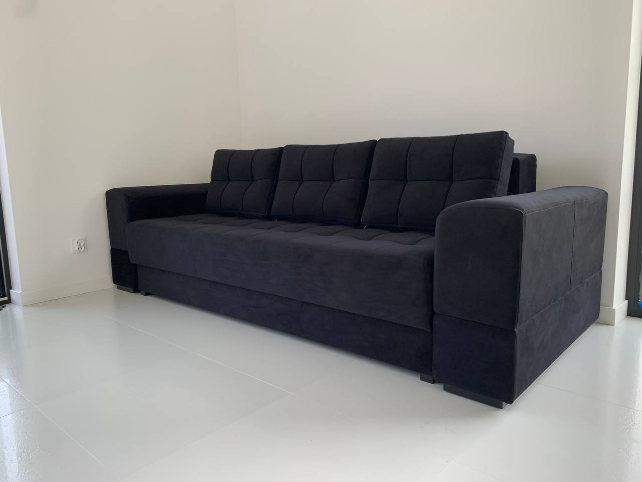 Czarna, rozkładana sofa z welurową tapicerką, ustawiona na białej podłodze w minimalistycznym wnętrzu. Widoczny fragment gniazdka elektrycznego.