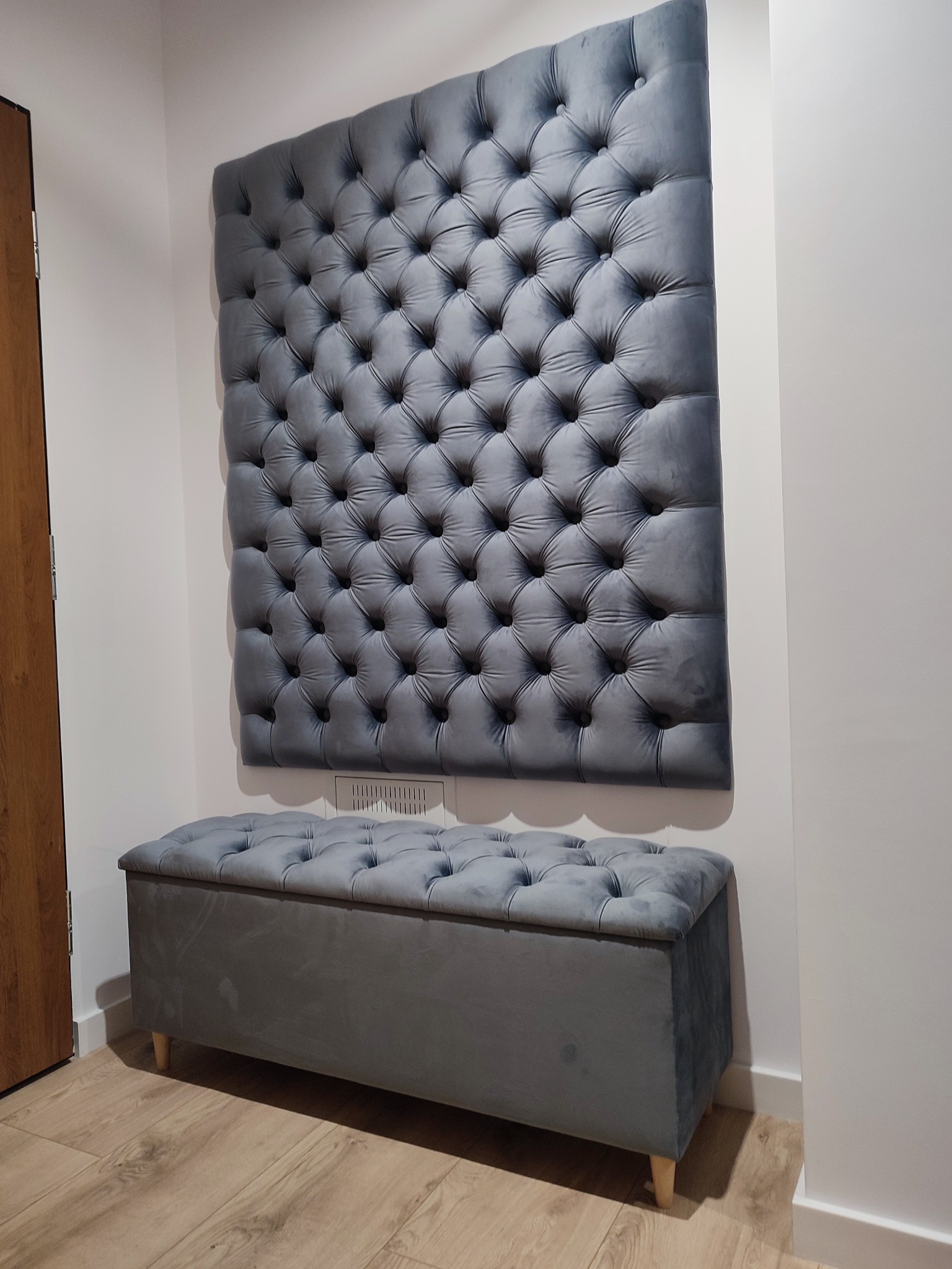 Szary, tapicerowany panel ścienny z pikowaniem i pufa ze schowkiem, ustawione w przedpokoju. Uwagę zwraca dopasowanie kolorystyczne i wzornicze obu elementów.