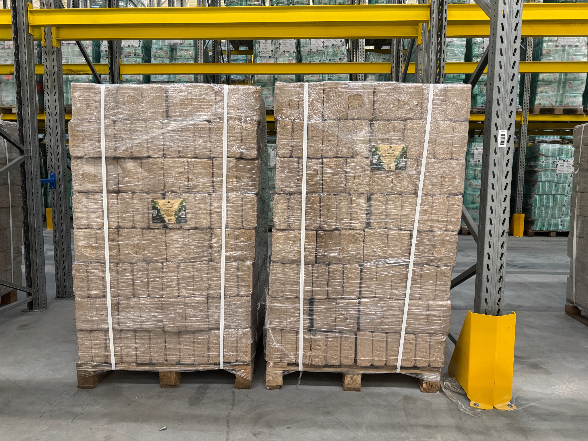 Dwie palety brykietu drzewnego RUF, owinięte folią, stoją w magazynie na tle regałów z towarem. Widoczny żółty odbojnik.