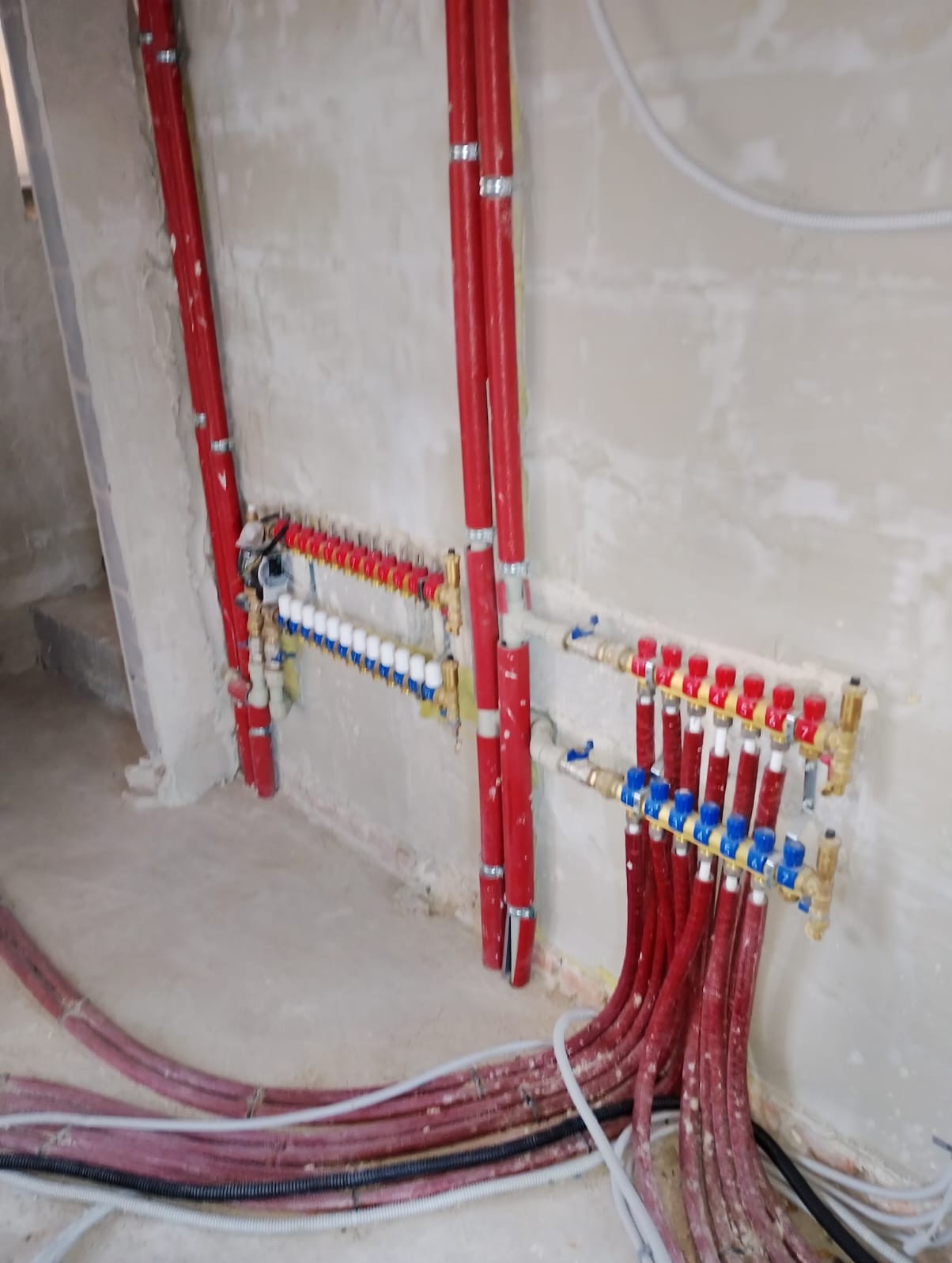 Rozdzielacz ogrzewania podłogowego z czerwonymi rurami w surowym wnętrzu. Widoczne zawory, rury doprowadzające i odprowadzające. Instalacja w trakcie montażu.
