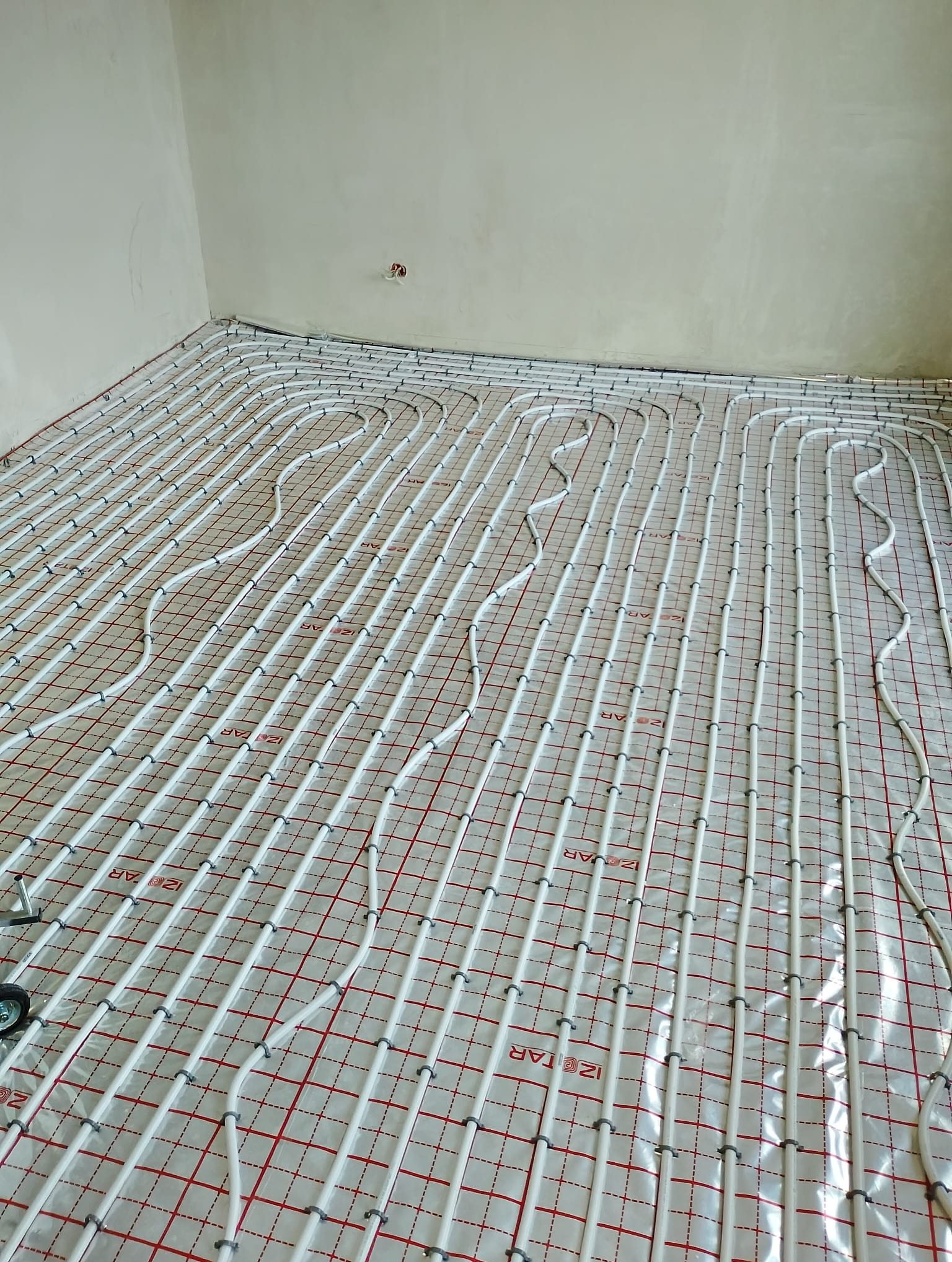 Białe rury ogrzewania podłogowego ułożone na folii z czerwoną siatką w surowym wnętrzu. Widok z góry, równoległe ułożenie rur, szare uchwyty mocujące.