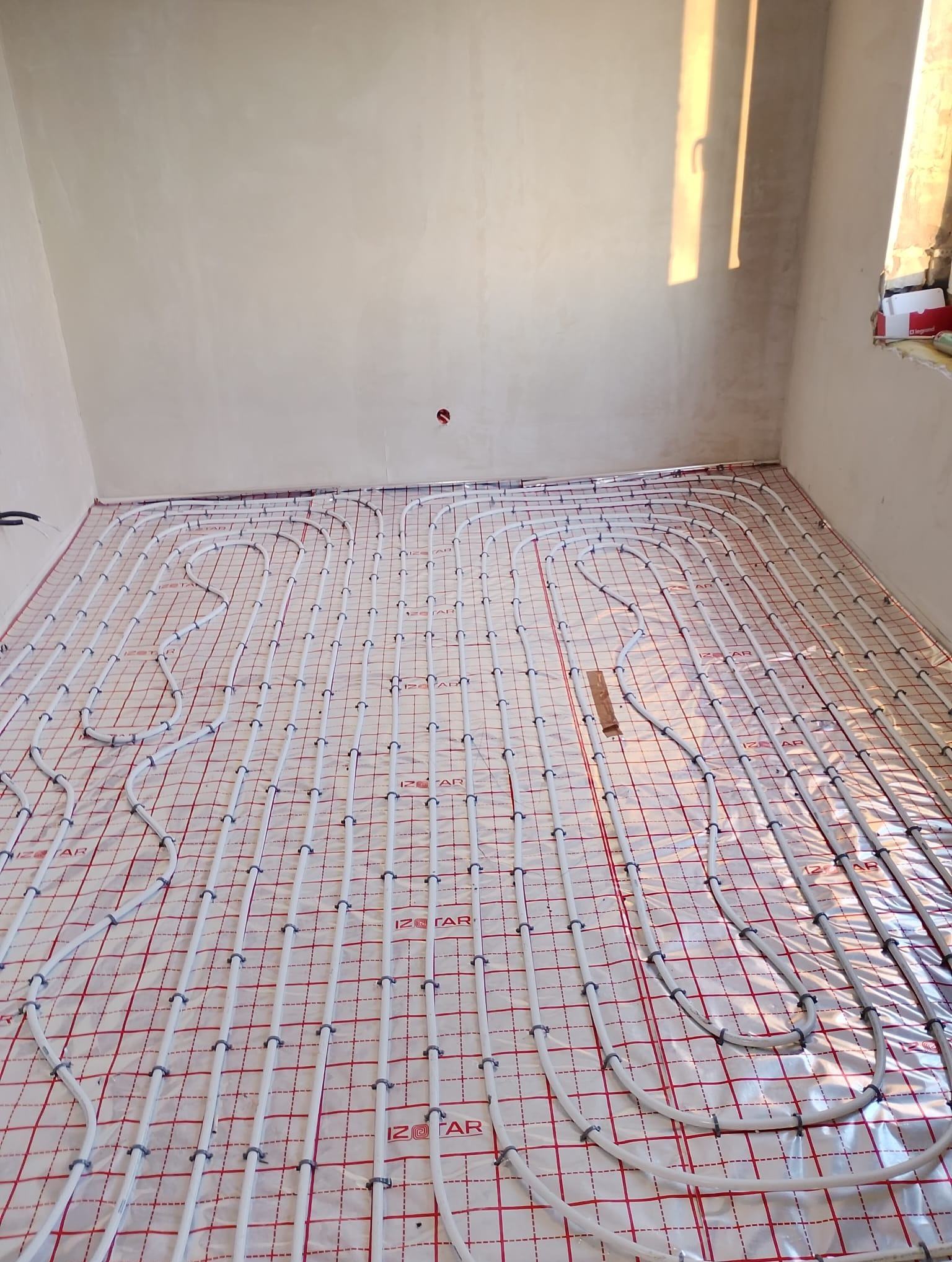Białe rury ogrzewania podłogowego ułożone na czerwonej macie w jasnym pomieszczeniu, widok z góry, rury przymocowane plastikowymi uchwytami, w tle okno.