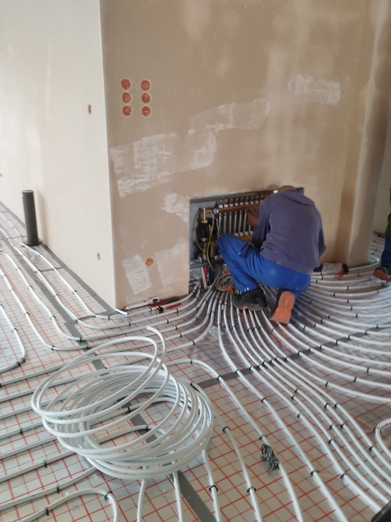 Instalacja ogrzewania podłogowego: białe rury ułożone na podłodze z czerwoną siatką, instalator w niebieskim kombinezonie podłącza rury do rozdzielacza.