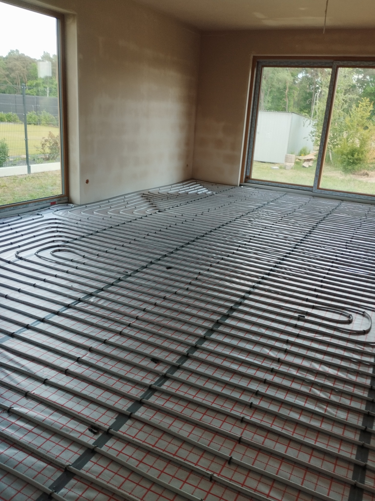 Ułożone rury ogrzewania podłogowego na izolacji z siatką w surowym wnętrzu z dużymi oknami, widok z góry. Etap instalacji przed wylewką.