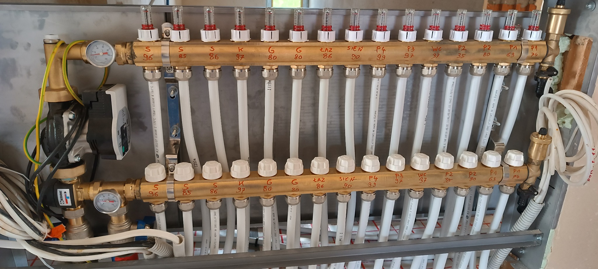 Rozdzielacz ogrzewania podłogowego z licznymi rurami, zaworami i manometrami, zamontowany na ścianie. Widoczne oznaczenia na elementach instalacji.