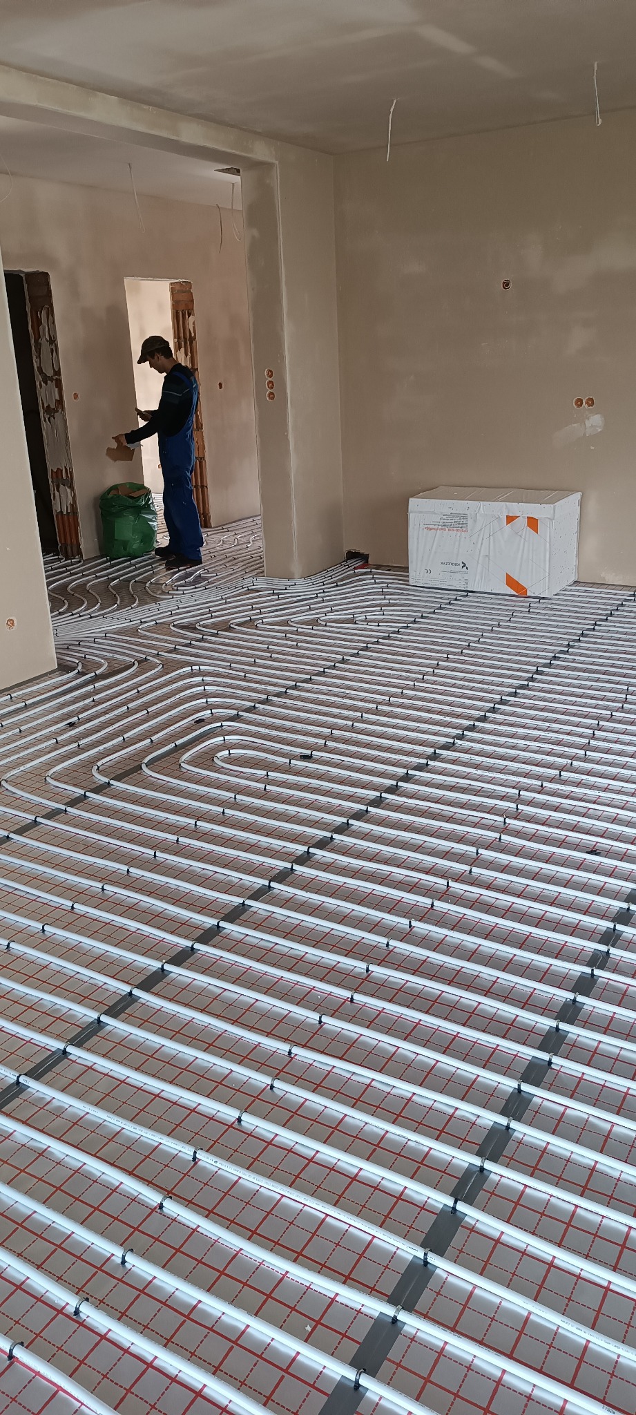 Białe rury ogrzewania podłogowego ułożone na czerwonej macie w jasnym pomieszczeniu. Instalator w kombinezonie w tle, obok zielony worek i biały karton.