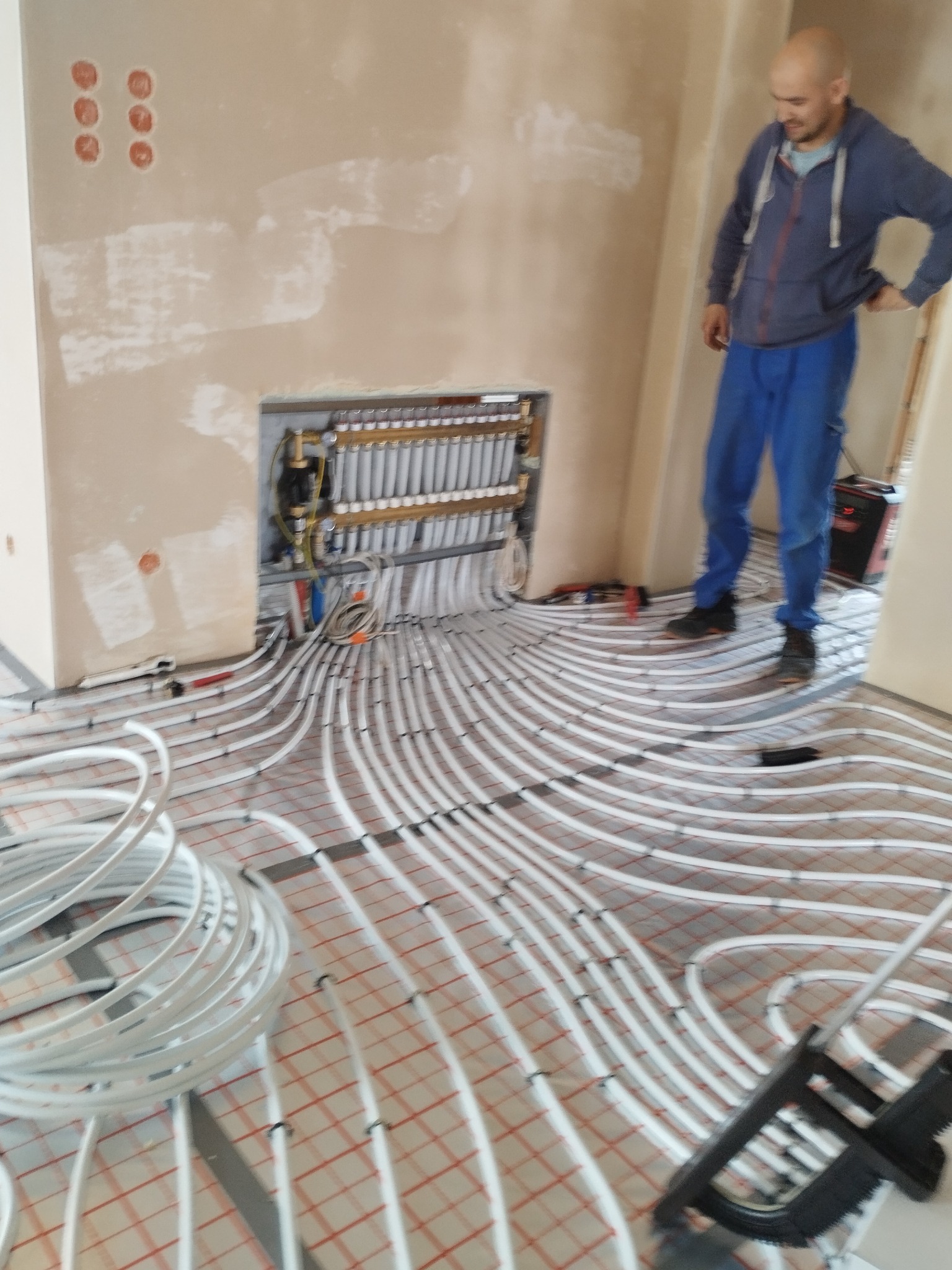 Białe rury ogrzewania podłogowego ułożone na czerwonej siatce, z rozdzielaczem w ścianie i instalatorem w tle. Widok na etapie montażu.