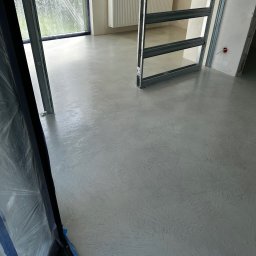 ALFA-BUD KM - Jasnoszara, wzorzysta posadzka w surowym wnętrzu z oknem zabezpieczonym folią i metalową konstrukcją drzwiową. Widoczne detale wykończenia ścian.