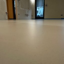 ALFA-BUD KM - Jasna, gładka posadzka w trakcie remontu, z widocznymi fragmentami ścian w stanie surowym i zabezpieczonymi oknami. Minimalistyczne wnętrze w trakcie wykończenia.