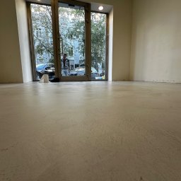 ALFA-BUD KM - Jasna, gładka posadzka w pustym pomieszczeniu z dużym oknem i widokiem na ulicę. Widoczny fragment ściany i sufitu. Minimalistyczny styl wykończenia.