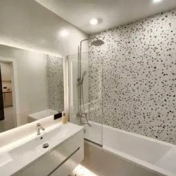 ALFA-BUD KM - Nowoczesna łazienka z wanną i kabiną prysznicową za szklaną ścianką. Umywalka z szafką i podświetlanym lustrem. Ściana wykończona wzorem lastryko.