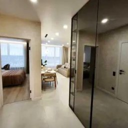 ALFA-BUD KM - Nowoczesne wnętrze mieszkania z lustrzaną szafą w przedpokoju, widok na salon i sypialnię. Jasne ściany i podłogi, minimalistyczny design.