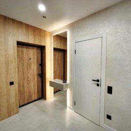 ALFA-BUD KM - Nowoczesny przedpokój z dwiema parami drzwi: drewnopodobne drzwi wejściowe i białe drzwi wewnętrzne z geometrycznym wzorem, lustro z podświetleniem i konsolą.