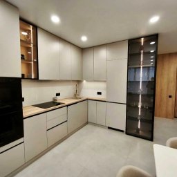 ALFA-BUD KM - Nowoczesna kuchnia w odcieniach szarości z drewnianym blatem, oświetleniem LED i przeszkloną witryną. Minimalistyczny design, widok na jadalnię i drewniane drzwi.