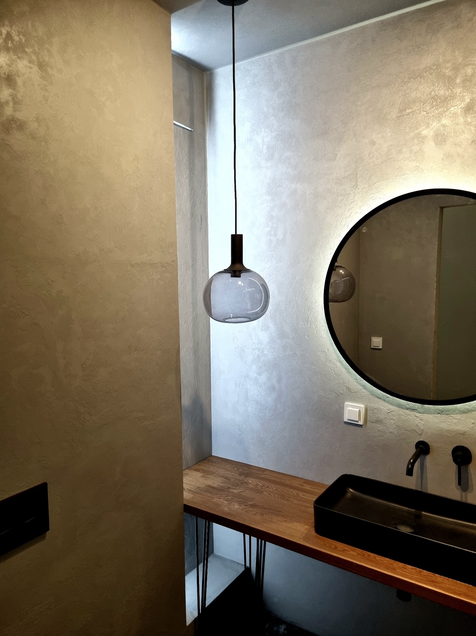 Stylowa łazienka z dekoracyjną ścianą w odcieniach szarości, okrągłym lustrem z podświetleniem LED i drewnianym blatem z czarną umywalką.