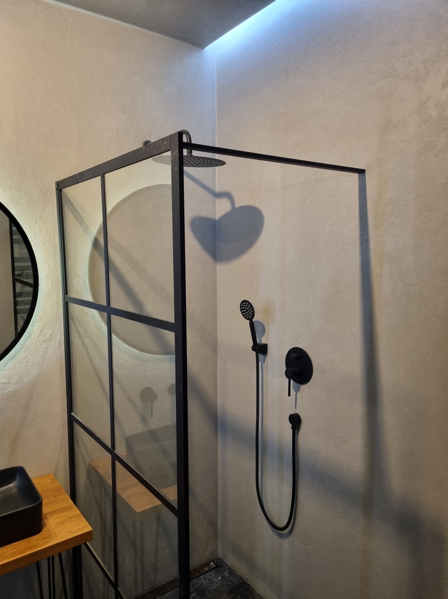 Nowoczesna kabina prysznicowa z czarną armaturą i szklaną ścianką działową na tle ściany wykończonej w odcieniach szarości, z elementami umywalki widocznymi po lewej stronie.