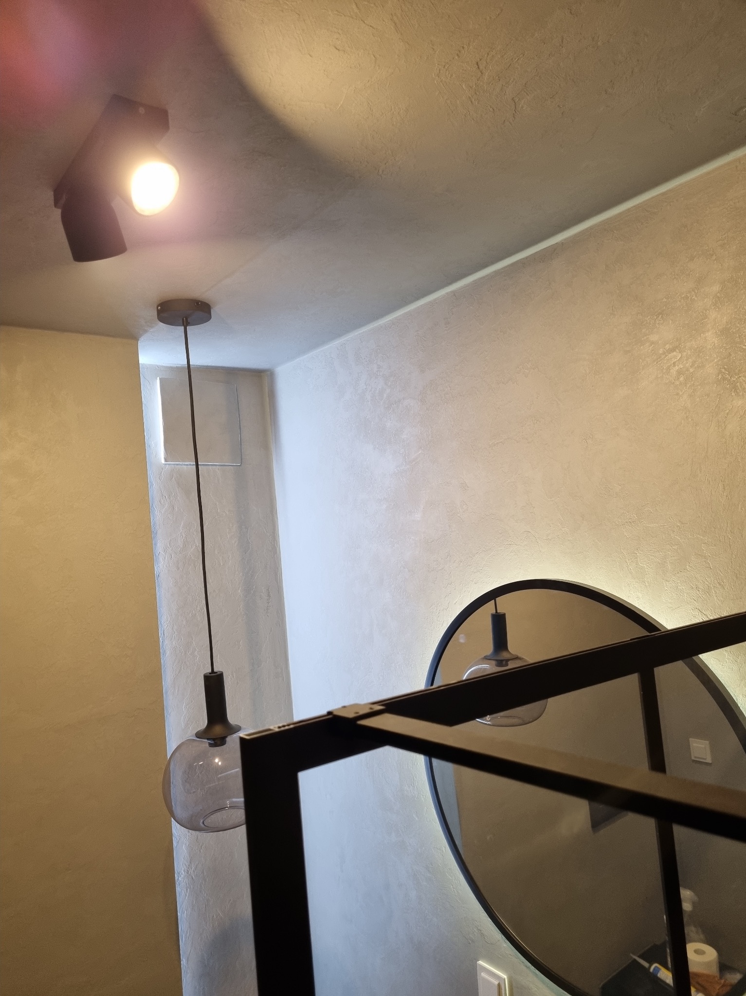 Nowoczesne oświetlenie sufitowe z lampą wiszącą i reflektorem punktowym, odbijające się w okrągłym lustrze z czarną ramą. Dekoracyjna faktura ściany.