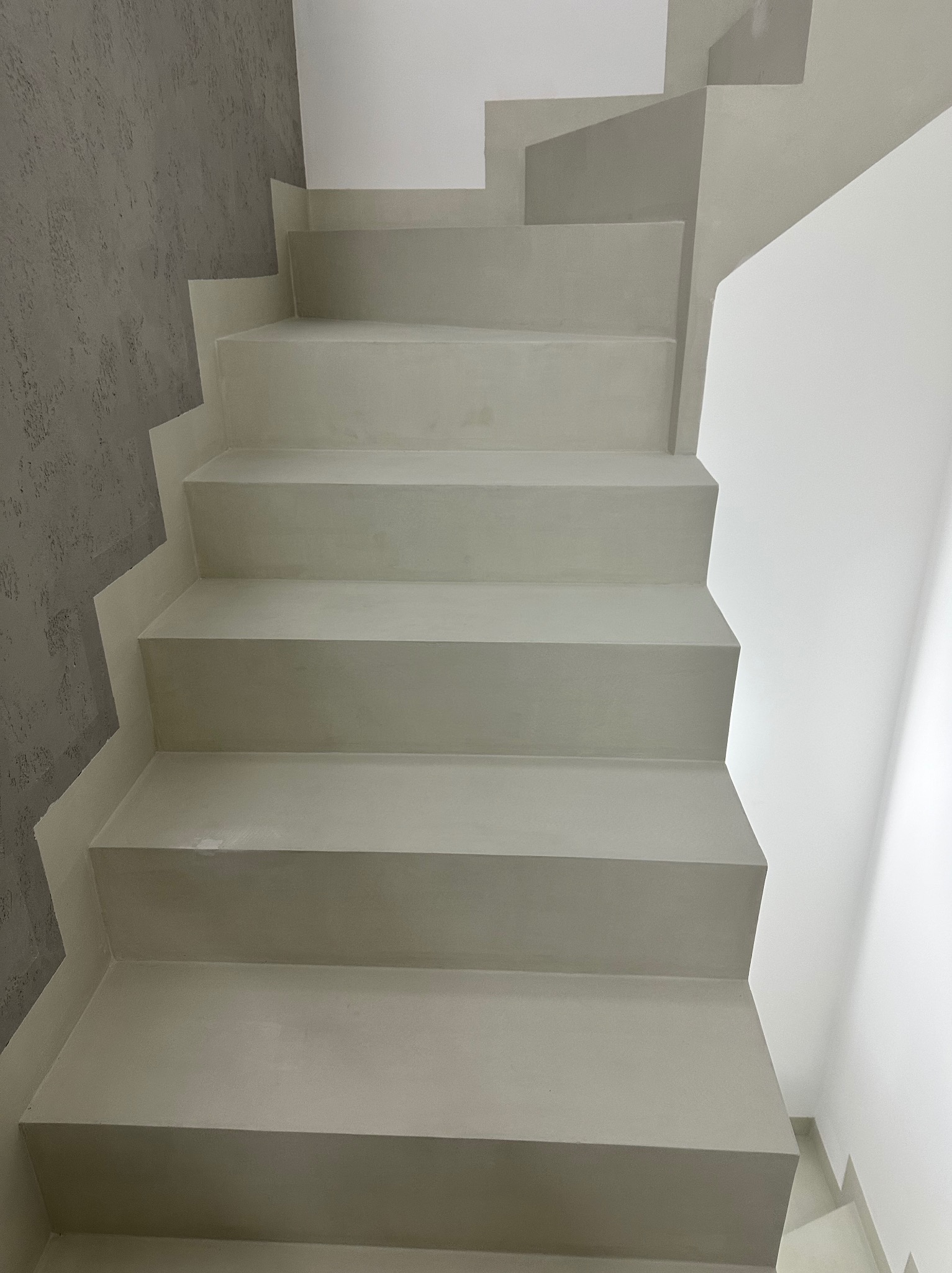 Wykończone schody wewnętrzne w odcieniach szarości, z dekoracyjną ścianą o fakturze betonu. Minimalistyczny design, widok z dołu, jasne oświetlenie.