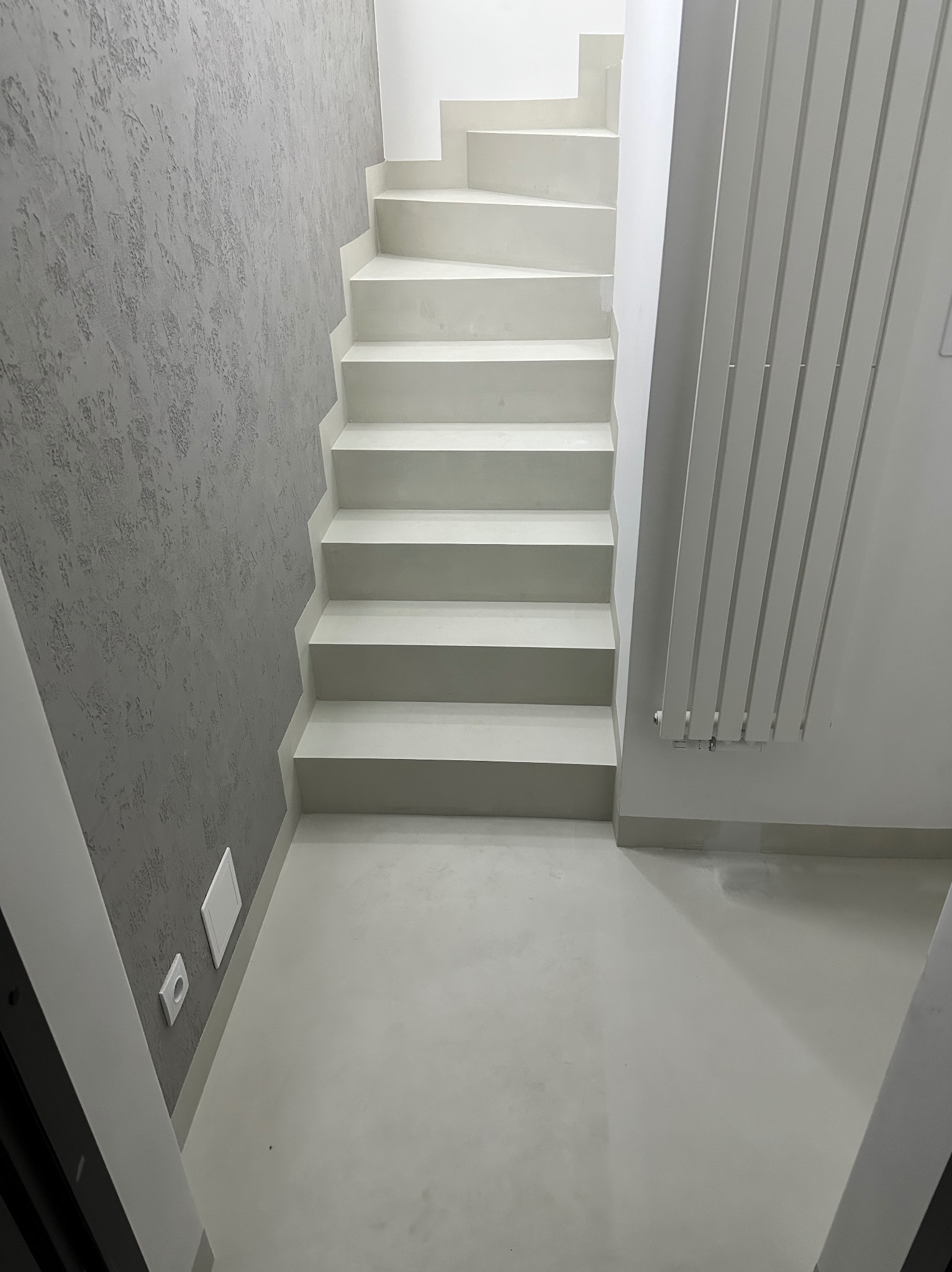 Minimalistyczne, jasne schody z mikrocementu w nowoczesnym wnętrzu. Szara ściana z dekoracyjną fakturą, biały grzejnik żeberkowy. Widok z dołu.