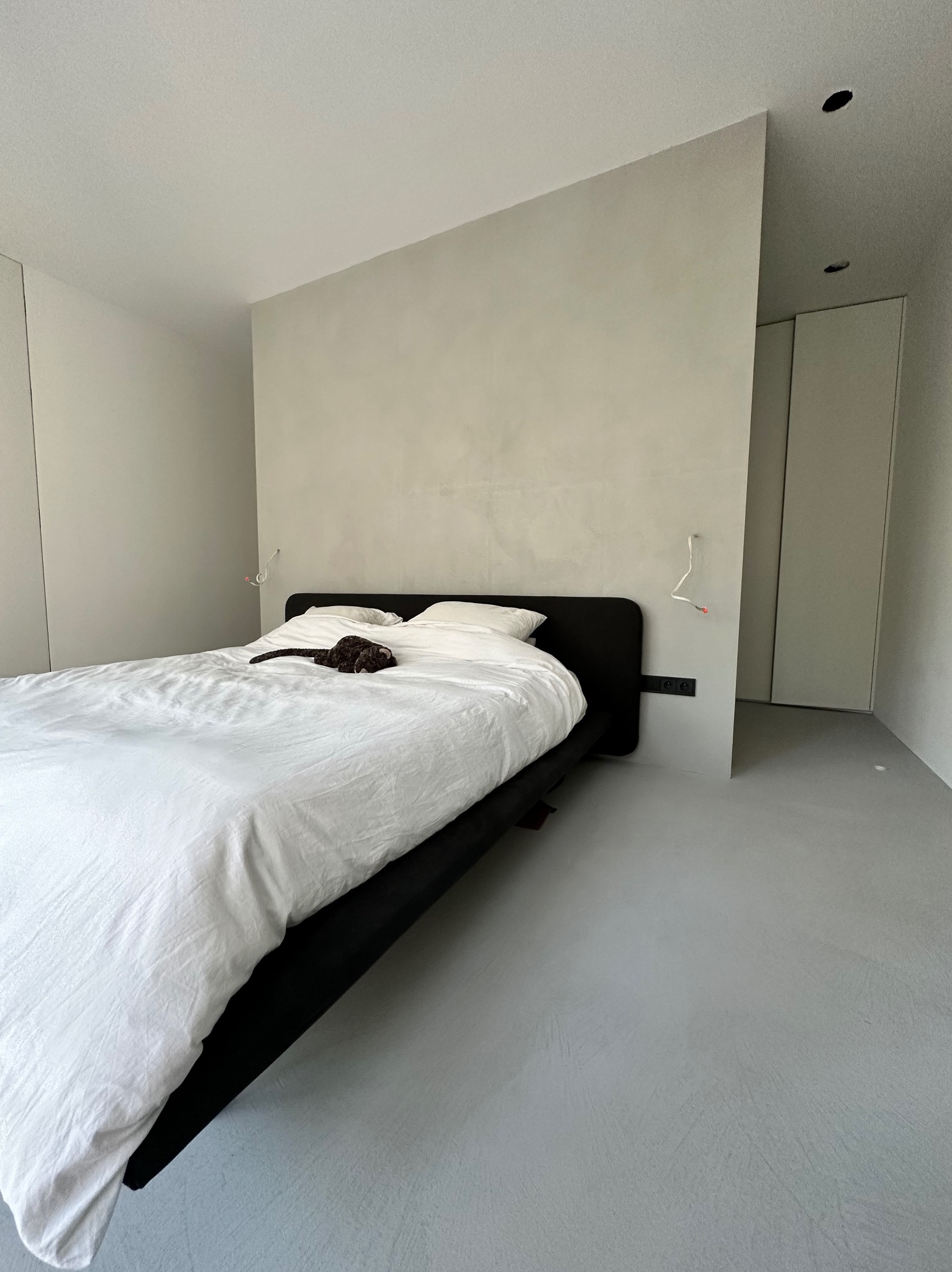 Minimalistyczna sypialnia z łóżkiem na tle ściany wykończonej mikrocementem i szarą posadzką. Widoczne gniazdka elektryczne i lampy ścienne.