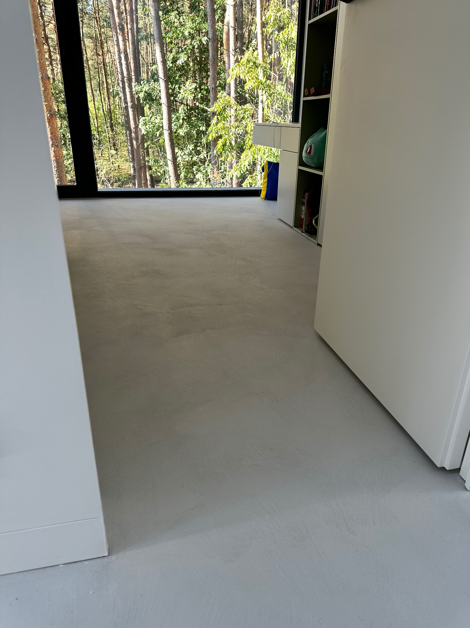 Jasnoszara posadzka mikrocementowa w nowoczesnym wnętrzu z dużym oknem na las. Minimalistyczny design, naturalne światło, widok na zieleń.