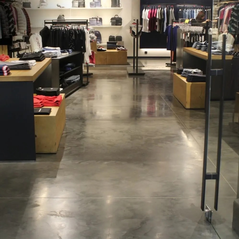 Polerowana posadzka betonowa w sklepie odzieżowym, odbijająca światło, z widocznymi elementami wyposażenia i odzieży w tle. Minimalistyczny design.