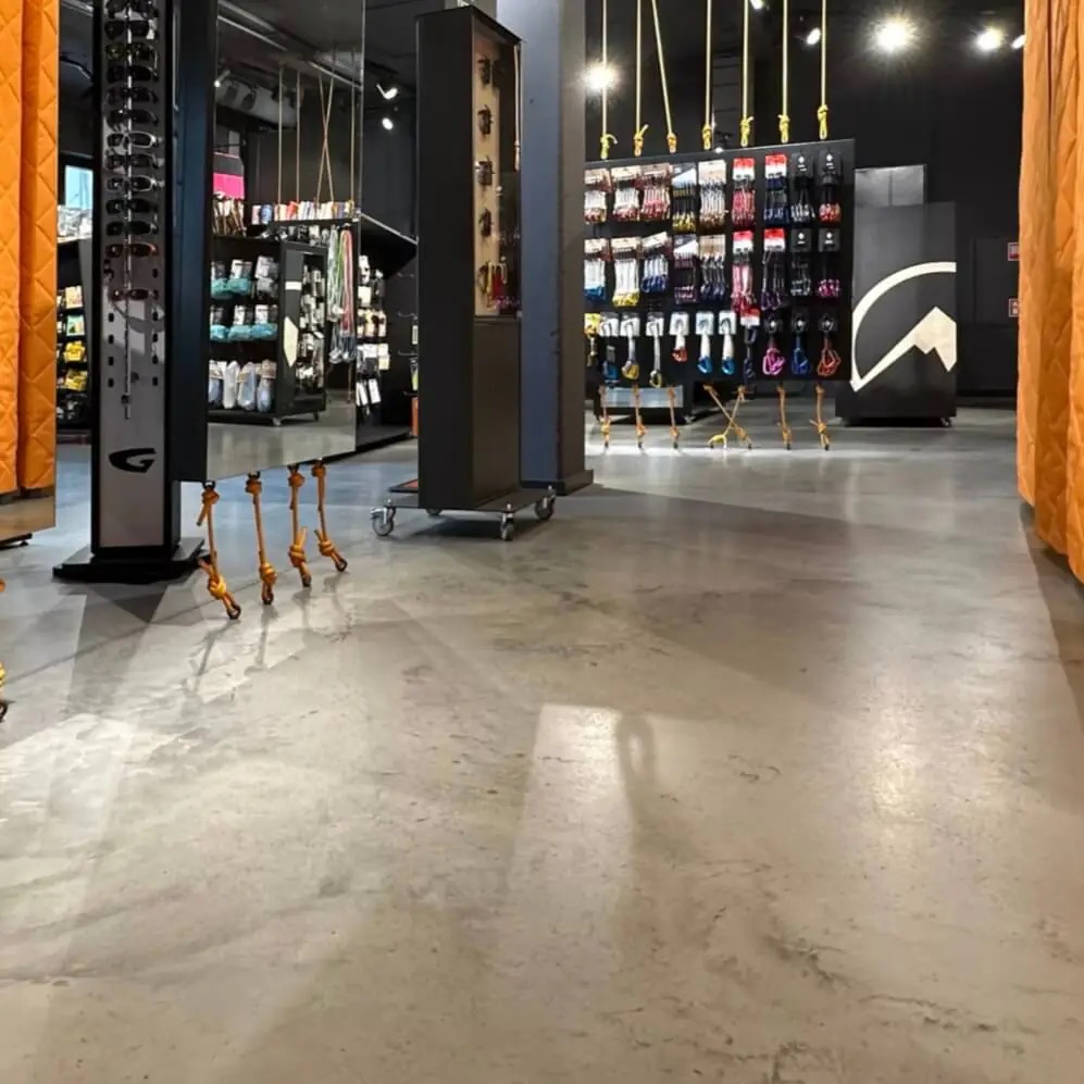 Jasnoszara posadzka w sklepie z akcesoriami wspinaczkowymi. Widoczne ekspozytory z produktami i oświetlenie sufitowe. Minimalistyczny design.