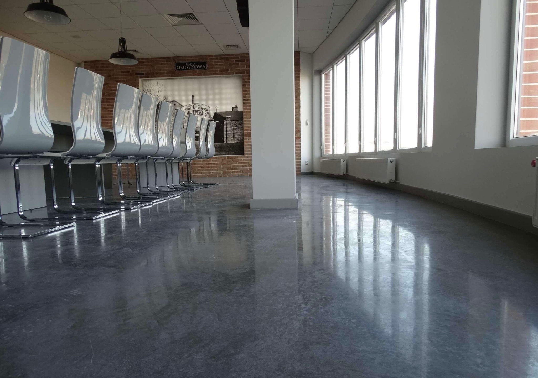 Błyszcząca, szara posadzka w przestronnym biurze z rzędem krzeseł i dużymi oknami. Widoczny refleks światła na gładkiej powierzchni podłogi.