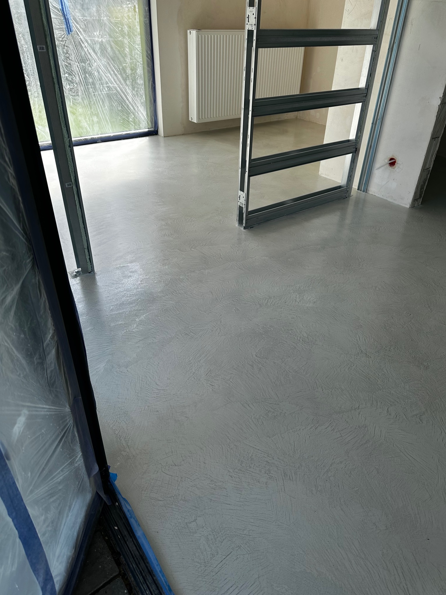 Jasnoszara, wzorzysta posadzka w surowym wnętrzu z oknem zabezpieczonym folią i metalową konstrukcją drzwiową. Widoczne detale wykończenia ścian.