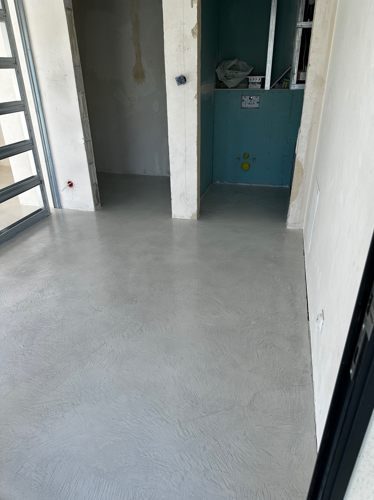 Szara, wzorzysta posadzka w surowym wnętrzu z widoczną instalacją sanitarną i elementami konstrukcyjnymi ścian. W tle widoczne płyty kartonowo-gipsowe.