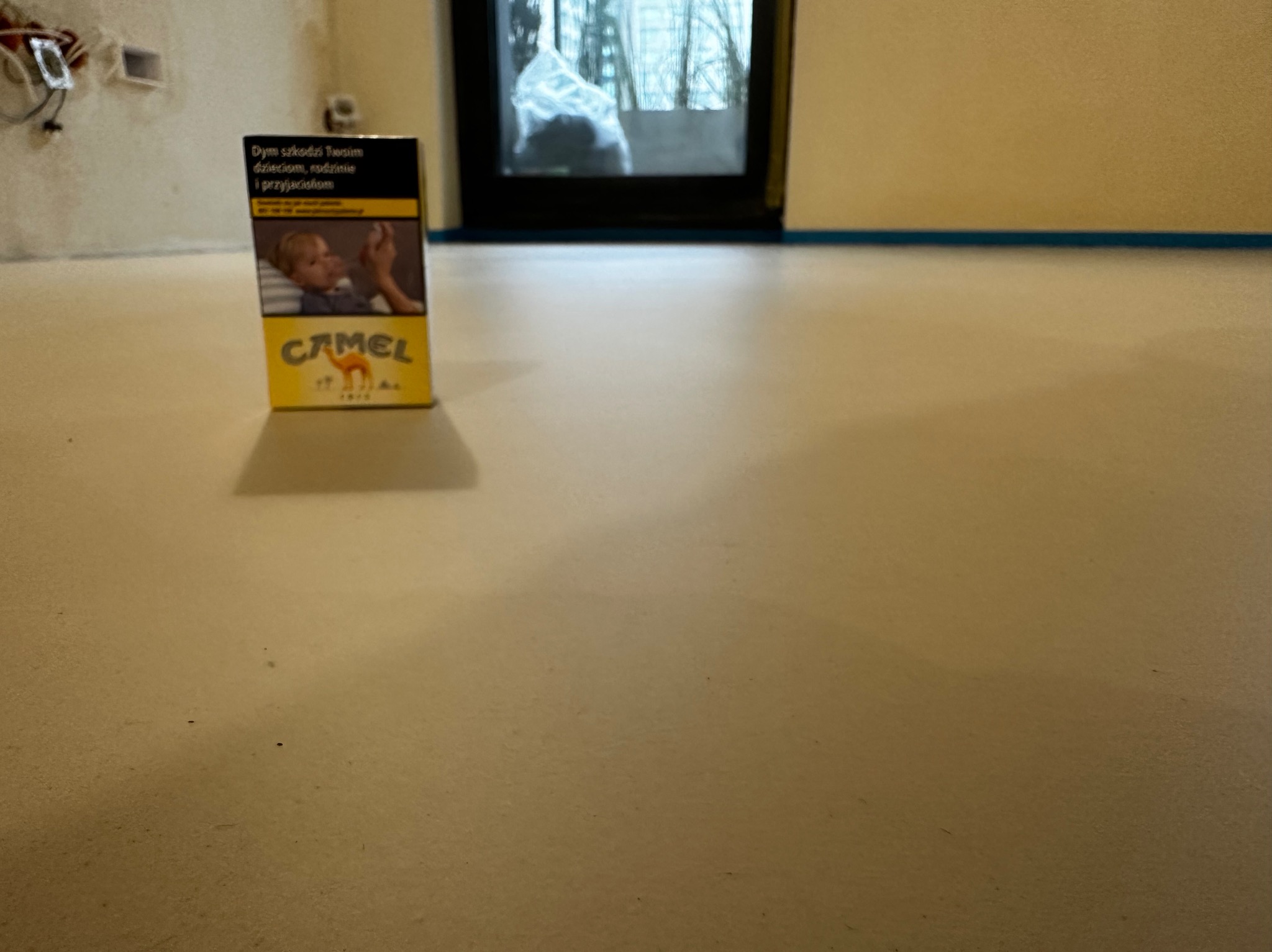 Jasna, gładka posadzka w pokoju z oknem, zabezpieczona taśmą malarską przy ścianach. Na pierwszym planie paczka papierosów Camel.
