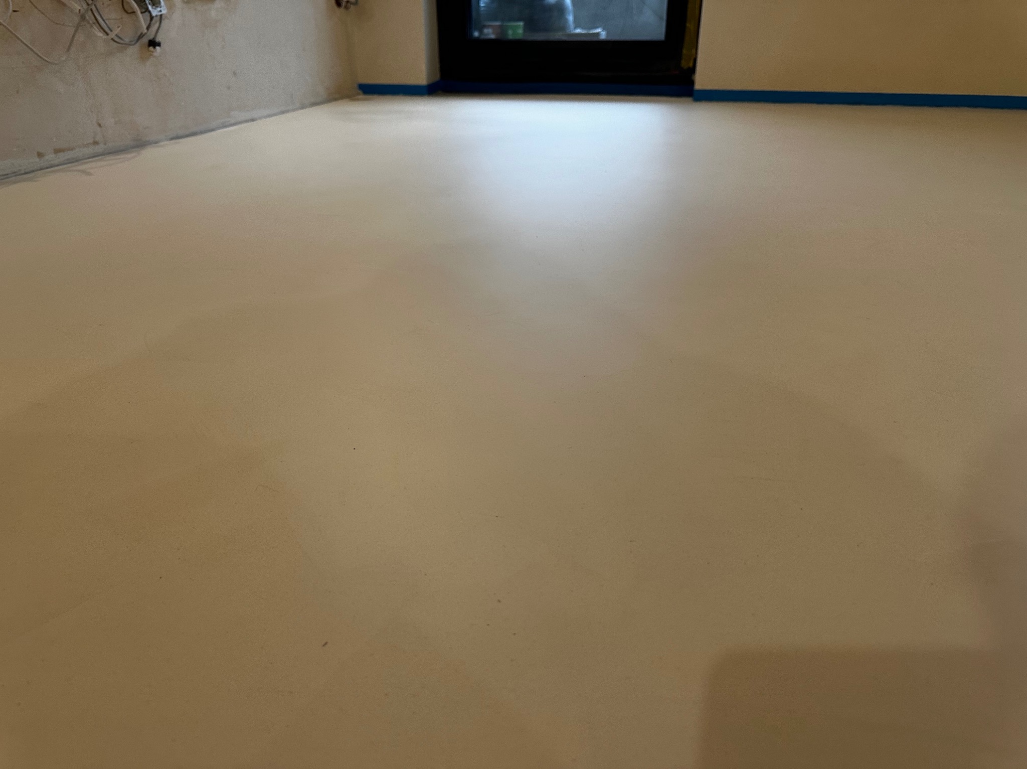 Jasna, gładka posadzka żywiczna w pomieszczeniu z widocznymi kablami przy ścianie i taśmą malarską zabezpieczającą krawędzie. Minimalistyczny design podłogi.