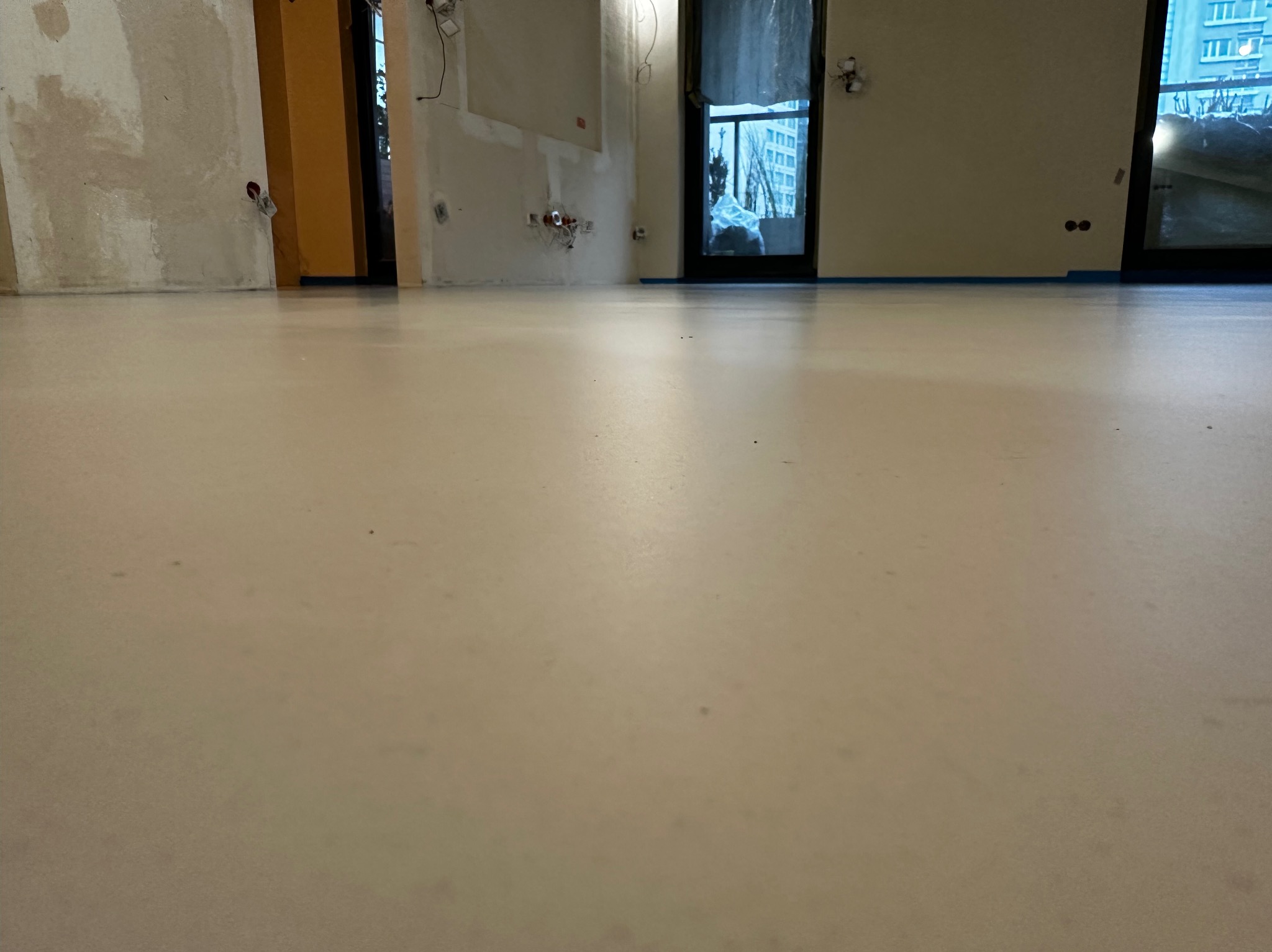 Jasna, gładka posadzka w trakcie remontu, z widocznymi fragmentami ścian w stanie surowym i zabezpieczonymi oknami. Minimalistyczne wnętrze w trakcie wykończenia.