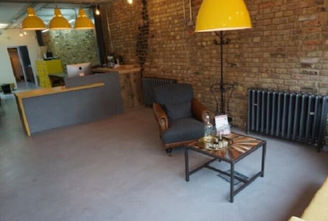 Jasnoszara posadzka w biurze z ceglaną ścianą, żółtymi lampami i ciemnym grzejnikiem. W tle recepcja i fotel z małym stolikiem. Widok na gładką, jednolitą powierzchnię.