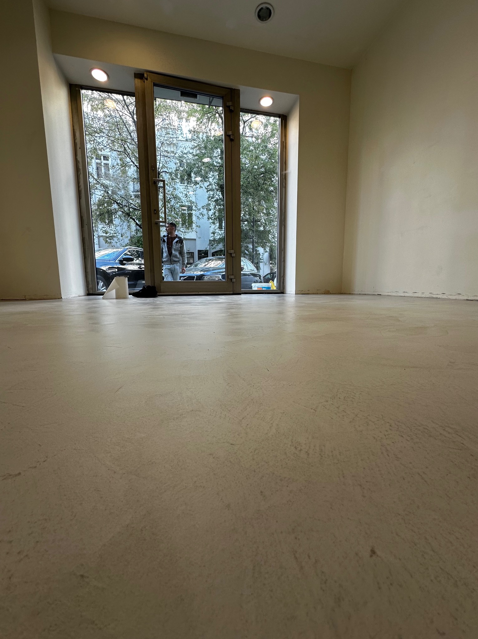 Jasna, gładka posadzka w pustym pomieszczeniu z dużym oknem i widokiem na ulicę. Widoczny fragment ściany i sufitu. Minimalistyczny styl wykończenia.