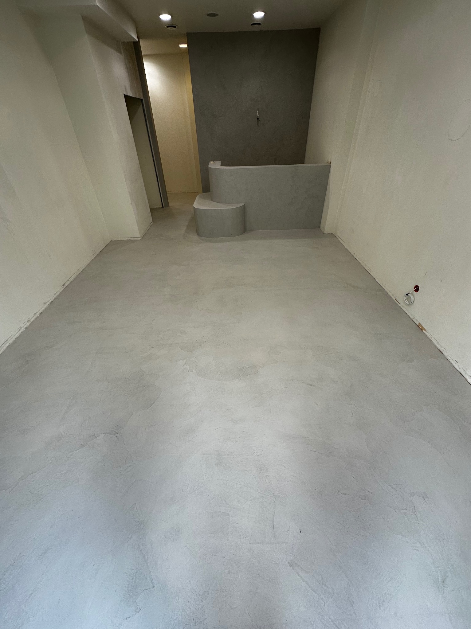 Jasnoszara posadzka z mikrocementu w pustym pomieszczeniu, widoczne ściany i fragment schodka. Minimalistyczne wykończenie wnętrza w neutralnej kolorystyce.