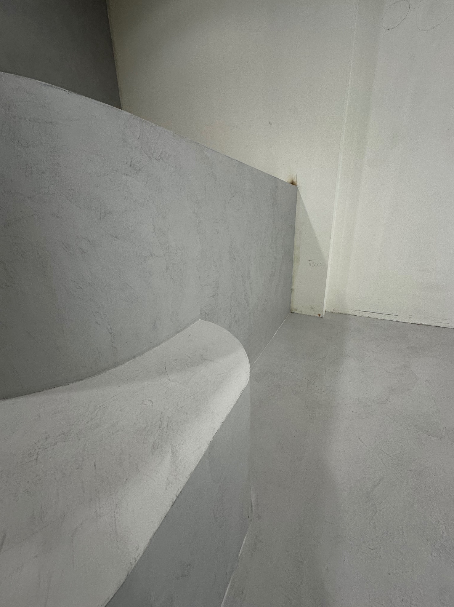 Szara, gładka powierzchnia ściany i podłogi z zaokrąglonym narożnikiem, wykończona w stylu minimalistycznym. Widoczne delikatne nierówności faktury.