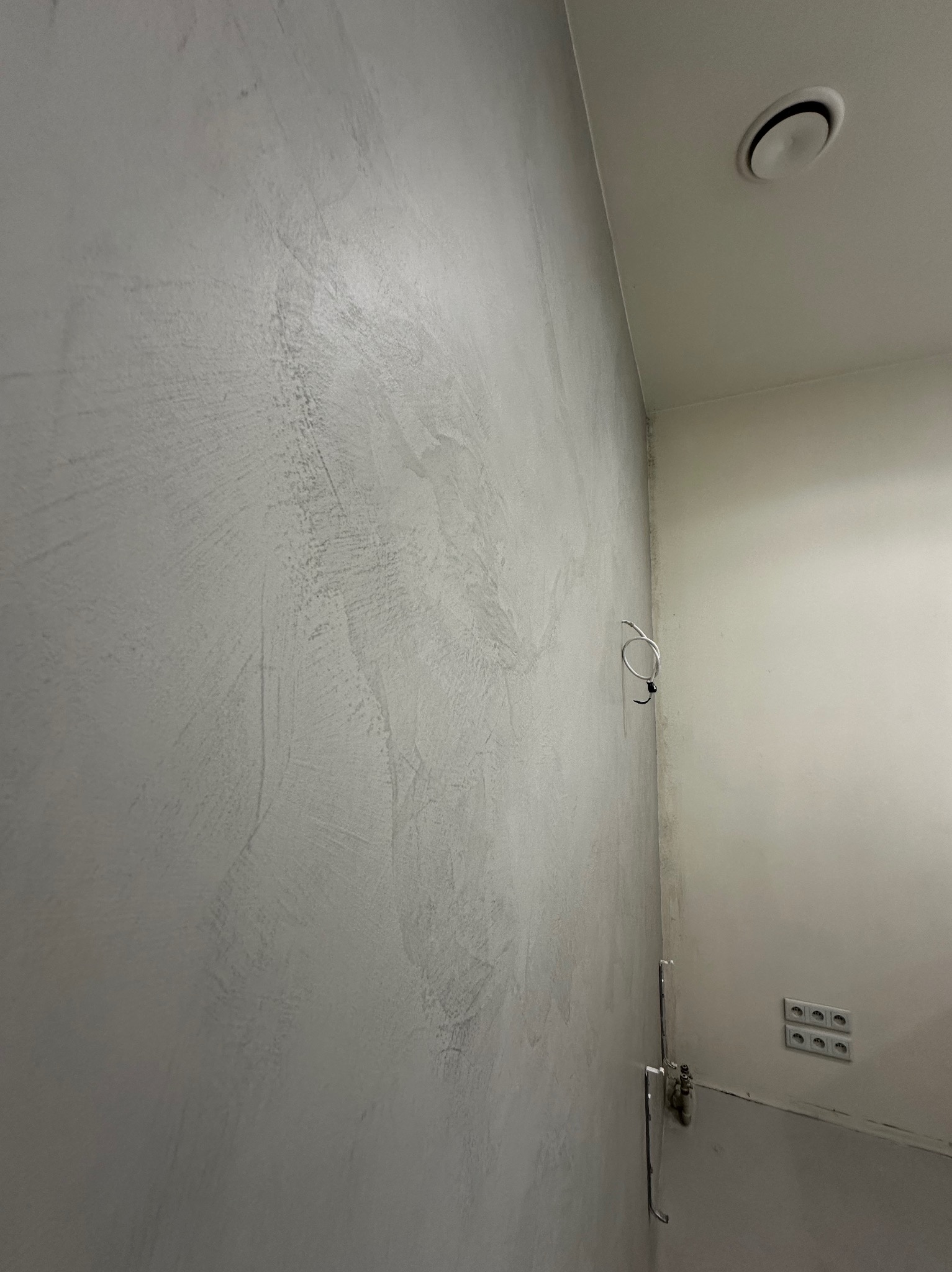 Ściana w łazience wykończona szarym mikrocementem z widocznymi detalami instalacji elektrycznej i wentylacji, ukazująca fakturę i minimalistyczny design.