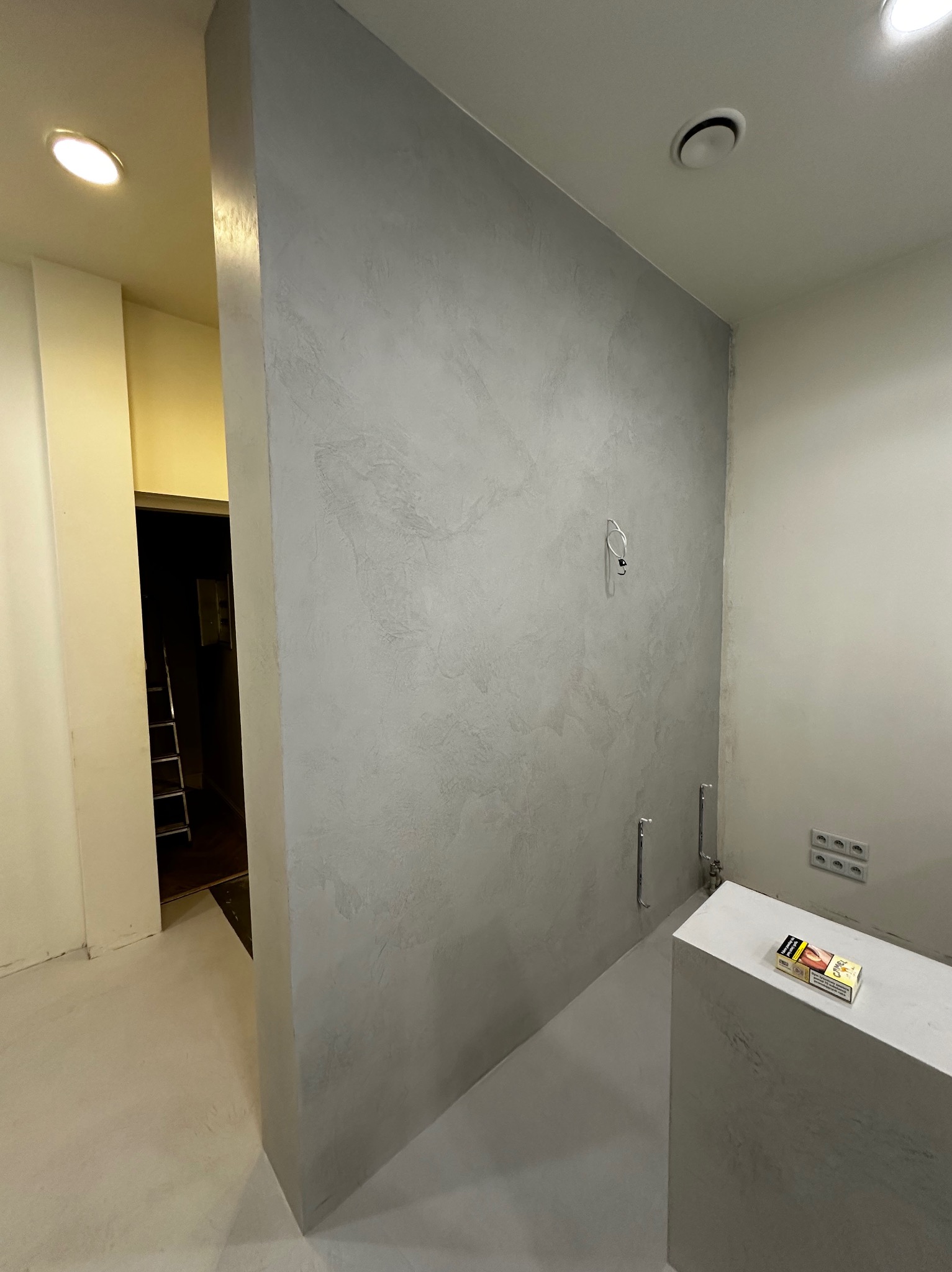 Ściana z efektem dekoracyjnym w odcieniach szarości, widoczne elementy instalacji. W tle fragment pomieszczenia z drabiną. Minimalistyczne wykończenie wnętrza.