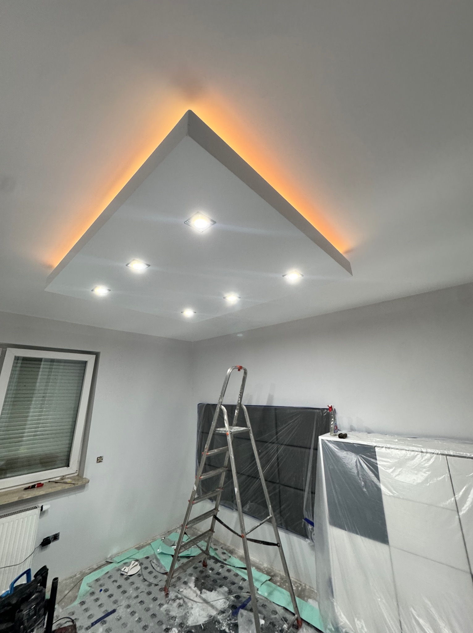 Wykończony sufit podwieszany z oświetleniem LED w kształcie trójkąta, widoczna drabina i zabezpieczone meble folią malarską. Świeżo pomalowane ściany w jasnym odcieniu.
