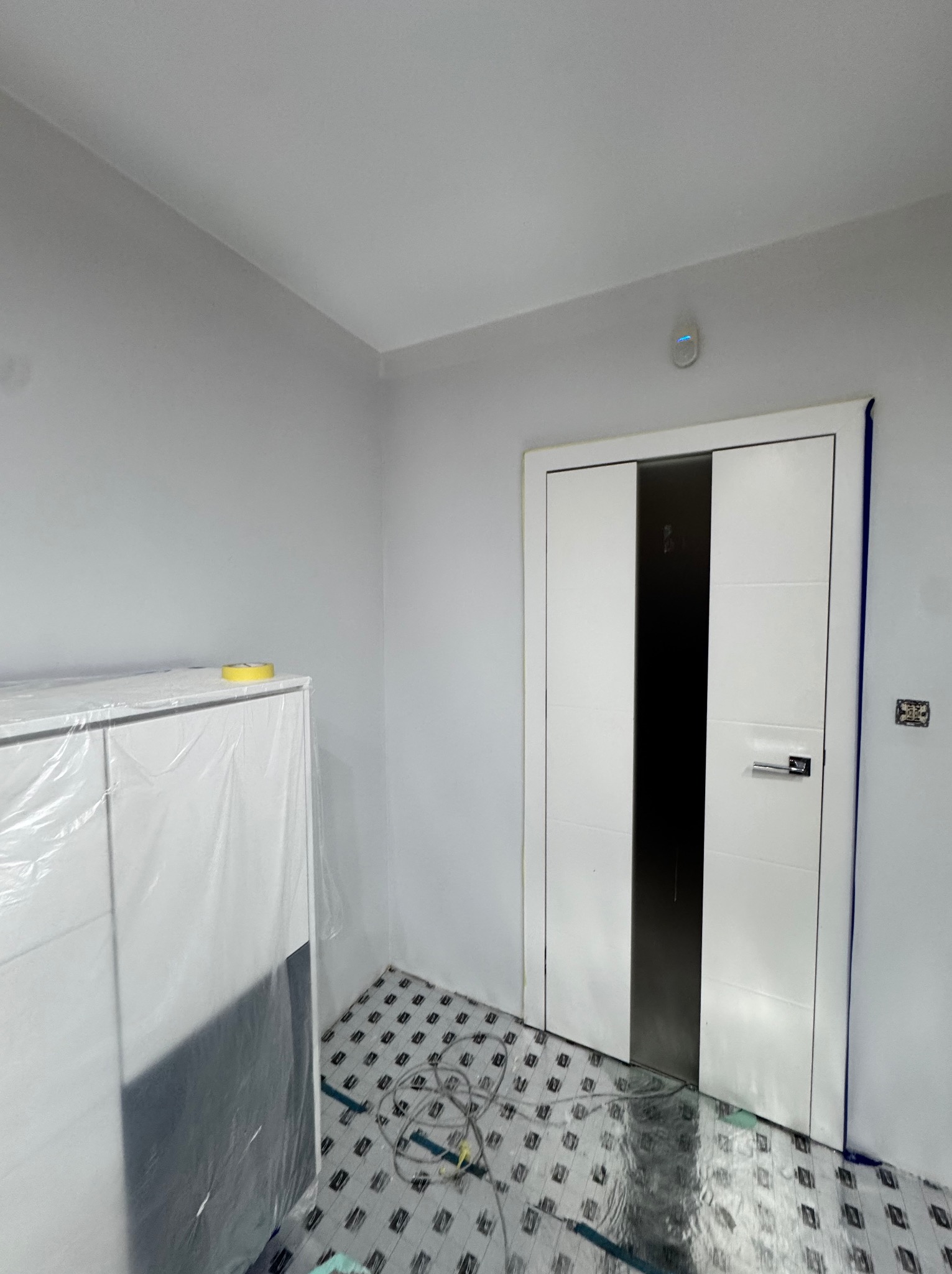 Białe drzwi z czarnym pasem w świeżo pomalowanym pokoju, zabezpieczone folią podłogi i meble, widoczny czujnik dymu na suficie.