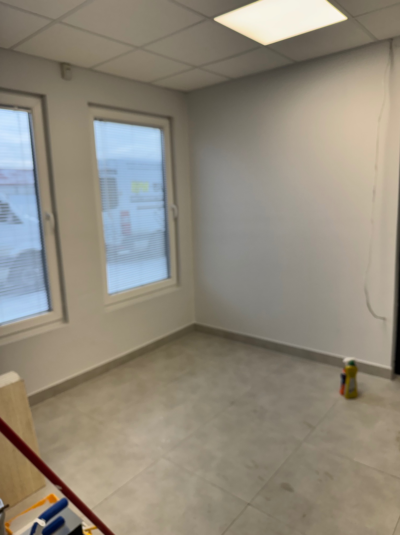 Pusty pokój biurowy w Lipnikach przed malowaniem. Widoczne dwa okna z żaluzjami, szare płytki na podłodze i narzędzia malarskie w rogu. Przygotowanie do prac wykończeniowych.