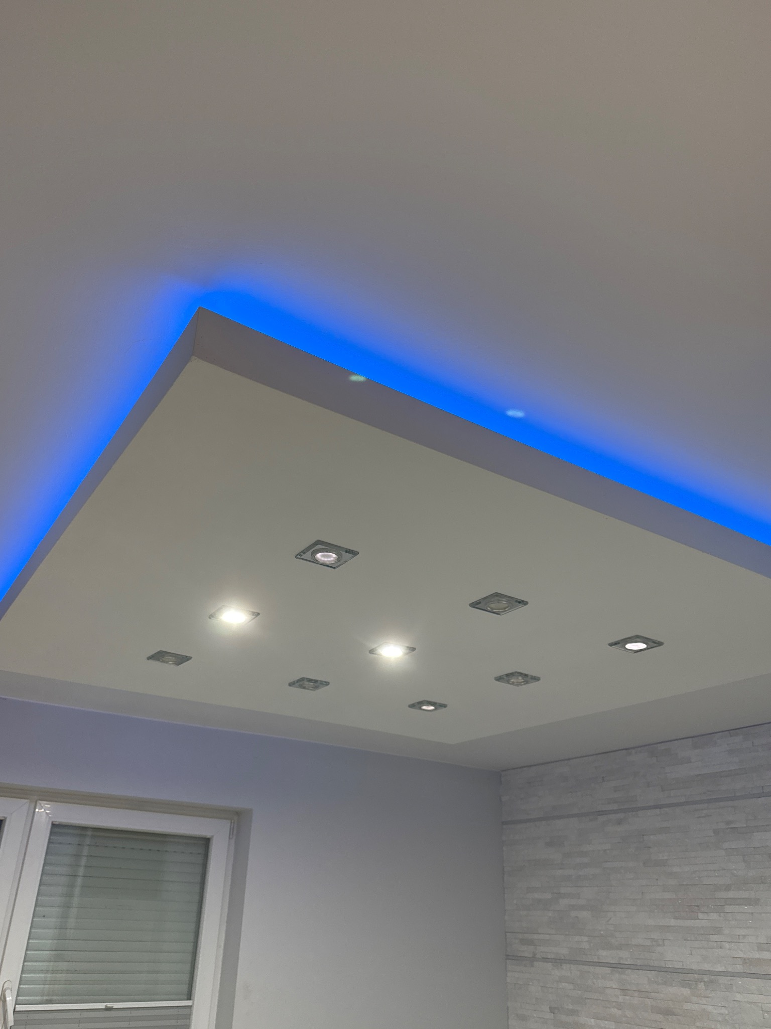 Podwieszany sufit z oświetleniem LED i punktowymi lampami, widok z dołu, fragment okna i ściany z dekoracyjną okładziną. Nowoczesne wykończenie wnętrza.