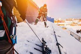 Pracownik w uprzęży odśnieża dach budynku szuflą, w tle widoczne inne budynki i błękitne niebo z promieniami słońca. Ujęcie z dołu, podkreślające stromy kąt dachu.