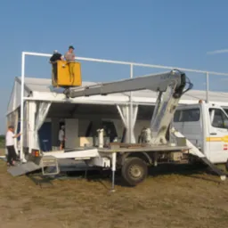 Montaż konstrukcji namiotowej z użyciem podnośnika koszowego; dwóch pracowników w żółtym koszu na tle białego namiotu i niebieskiego nieba.