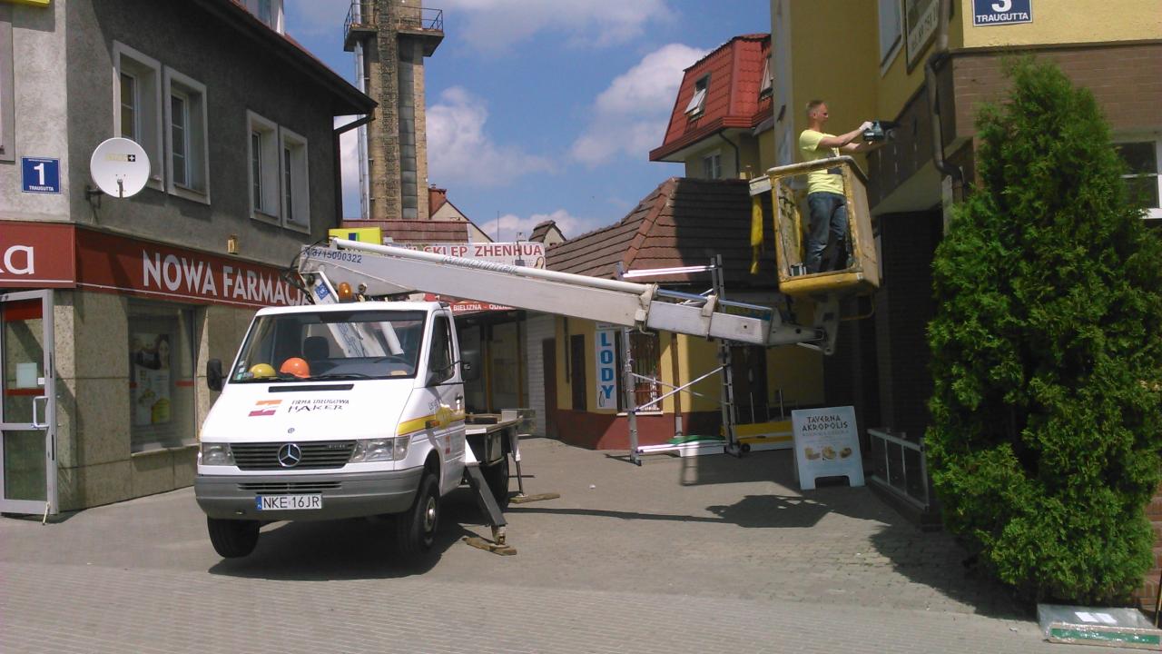Podnośnik koszowy na chodniku przed budynkiem. Pracownik w koszu podnośnika wykonuje prace na wysokości. Widoczny budynek apteki i restauracji.
