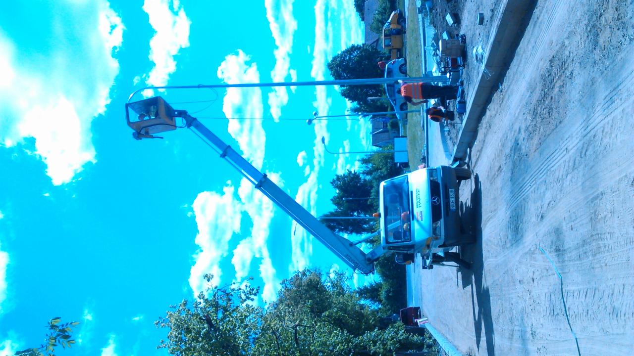 Podnośnik koszowy z wysięgnikiem podczas prac na zewnątrz, operator w koszu, pracownik w pomarańczowej kamizelce przy chodniku. Błękitne niebo w tle.