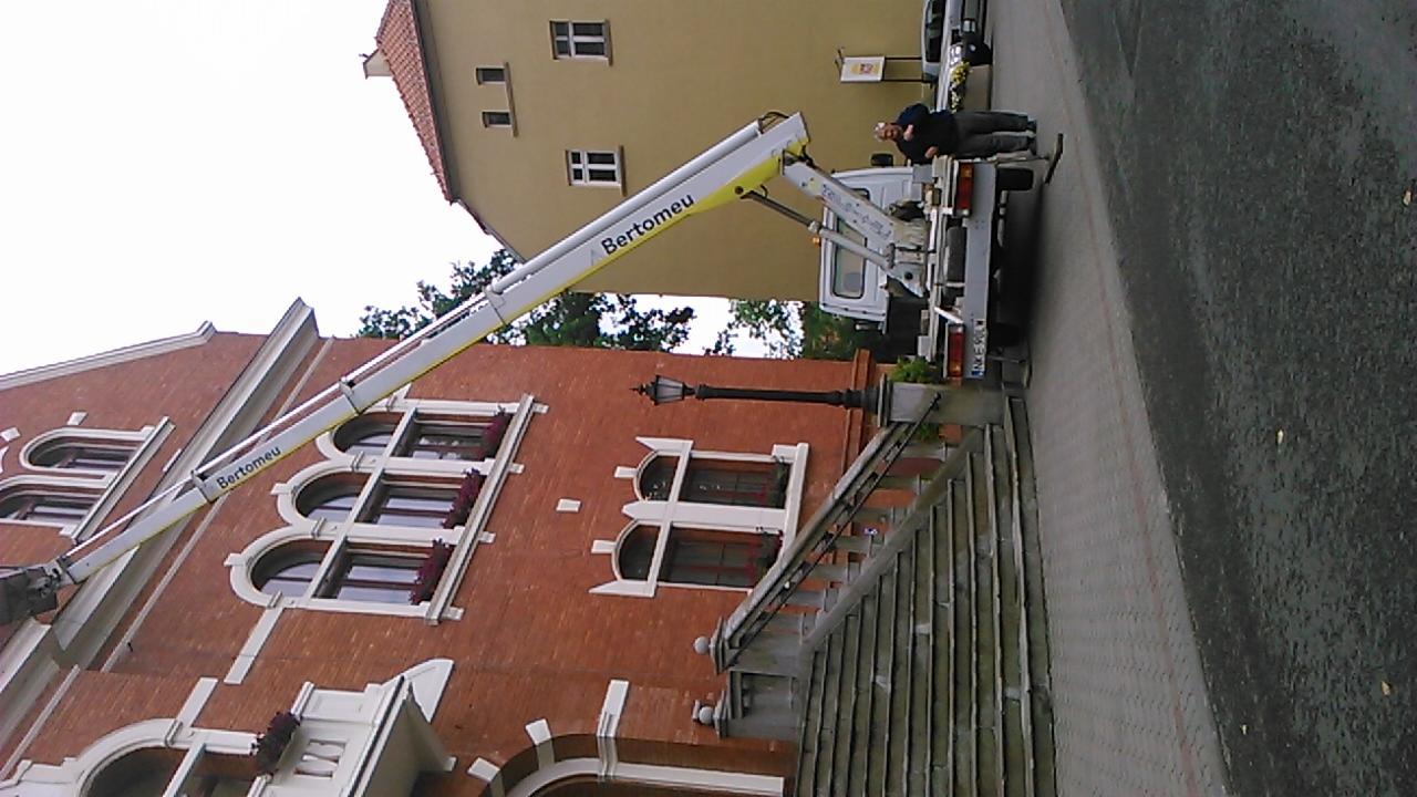 Podnośnik koszowy 'Bertomeu' przy budynku z czerwonej cegły. Operator na platformie, widoczne schody i ulica. Ujęcie z dołu, obrócone o 90 stopni w prawo.